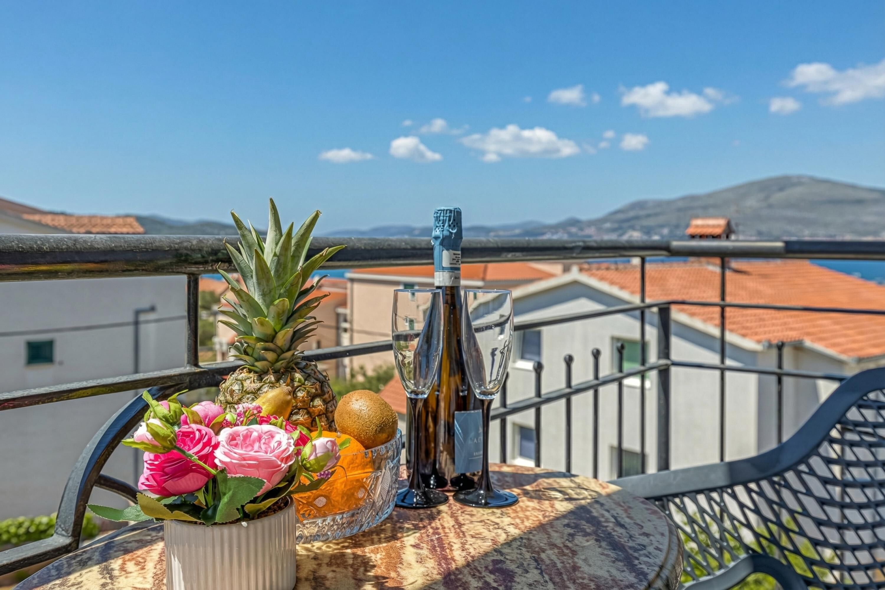 Balcón con una mesa que sostiene una piña, flores y una botella, con vistas a una zona costera con edificios y una montaña.