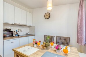 Une cuisine moderne avec des armoires blanches, un four à micro-ondes et un petit réfrigérateur. On y trouve une table à manger avec un set de table, une horloge au mur et un rideau rose.