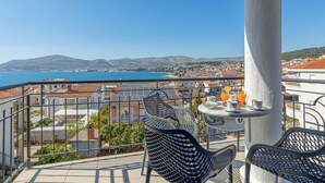 Ein Balkon mit Tisch und Stühlen, mit Blick auf eine Küstenstadt, Gebäude und das klare, blaue Meer.