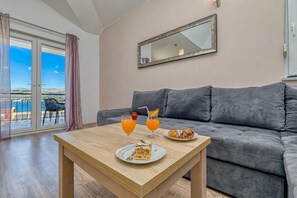 Ein modernes Wohnzimmer mit einer grauen Couch, einem Holztisch mit Speisen und Getränken sowie einem Balkon mit Blick.