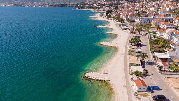 Ein Luftbild einer Küstenstadt mit einem langen, schmalen Strand, klarem, blauem Wasser und Wohngebäuden entlang der Küste.