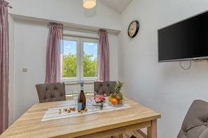 A dining area with a wooden table, two chairs, a bottle of wine, glasses, a fruit bowl, and a clock on the wall.