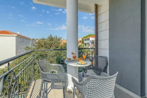 A balcony with two wicker chairs, a small table with food and drinks, and a view of buildings and trees.
