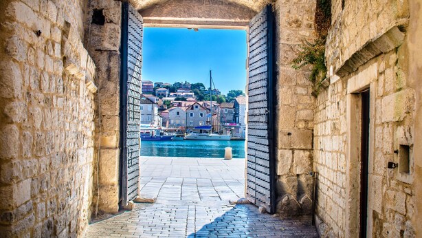 A stone archway leading to a harbor with buildings and boats.