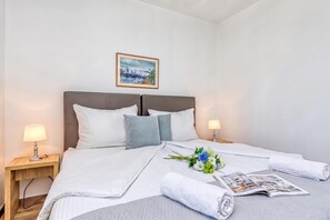 Ein ordentlich bezogenes Bett mit weißer Daunendecke, Kissen und einer Zeitschrift. Zwei Nachttische mit Lampen und ein gerahmtes Bild an der Wand über dem Bett.