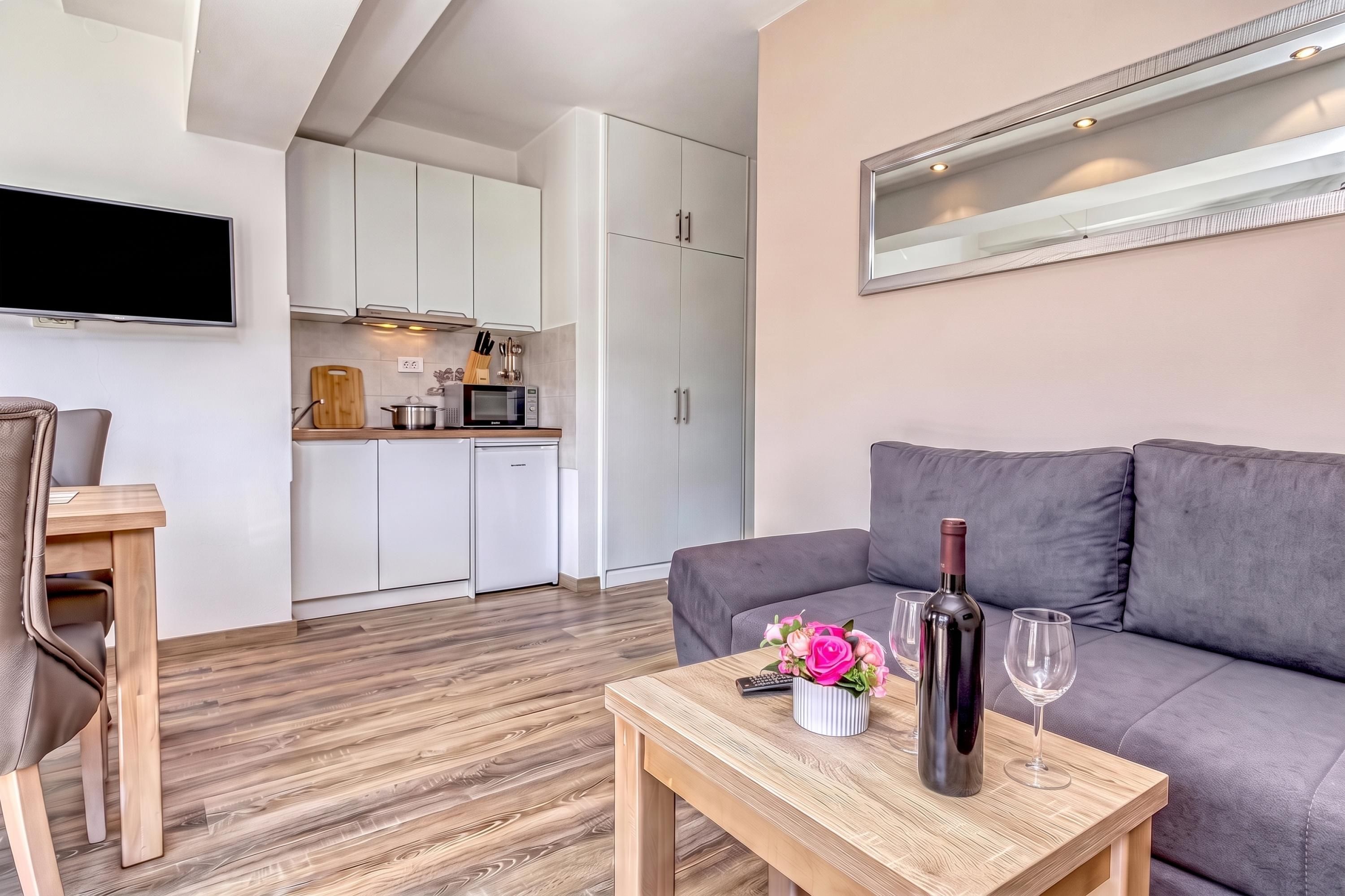 A modern living room with a grey sofa, a wooden coffee table with a bottle of wine and glasses, and a TV mounted on the wall.