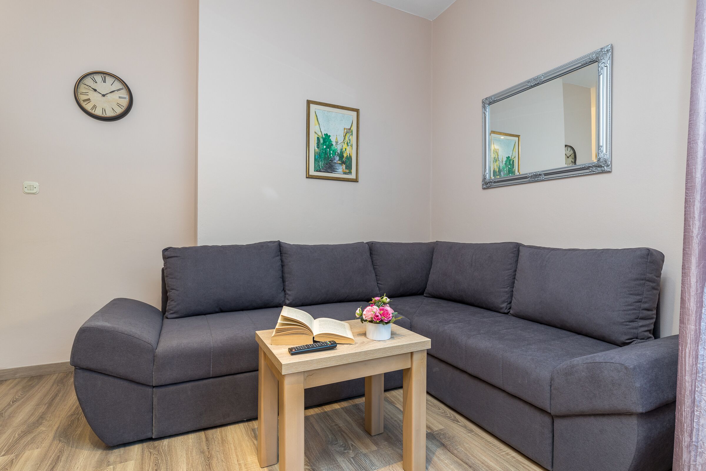 A modern living room with a gray sofa, a wooden coffee table with an open book and a vase of flowers, a clock, and a framed picture on the wall.