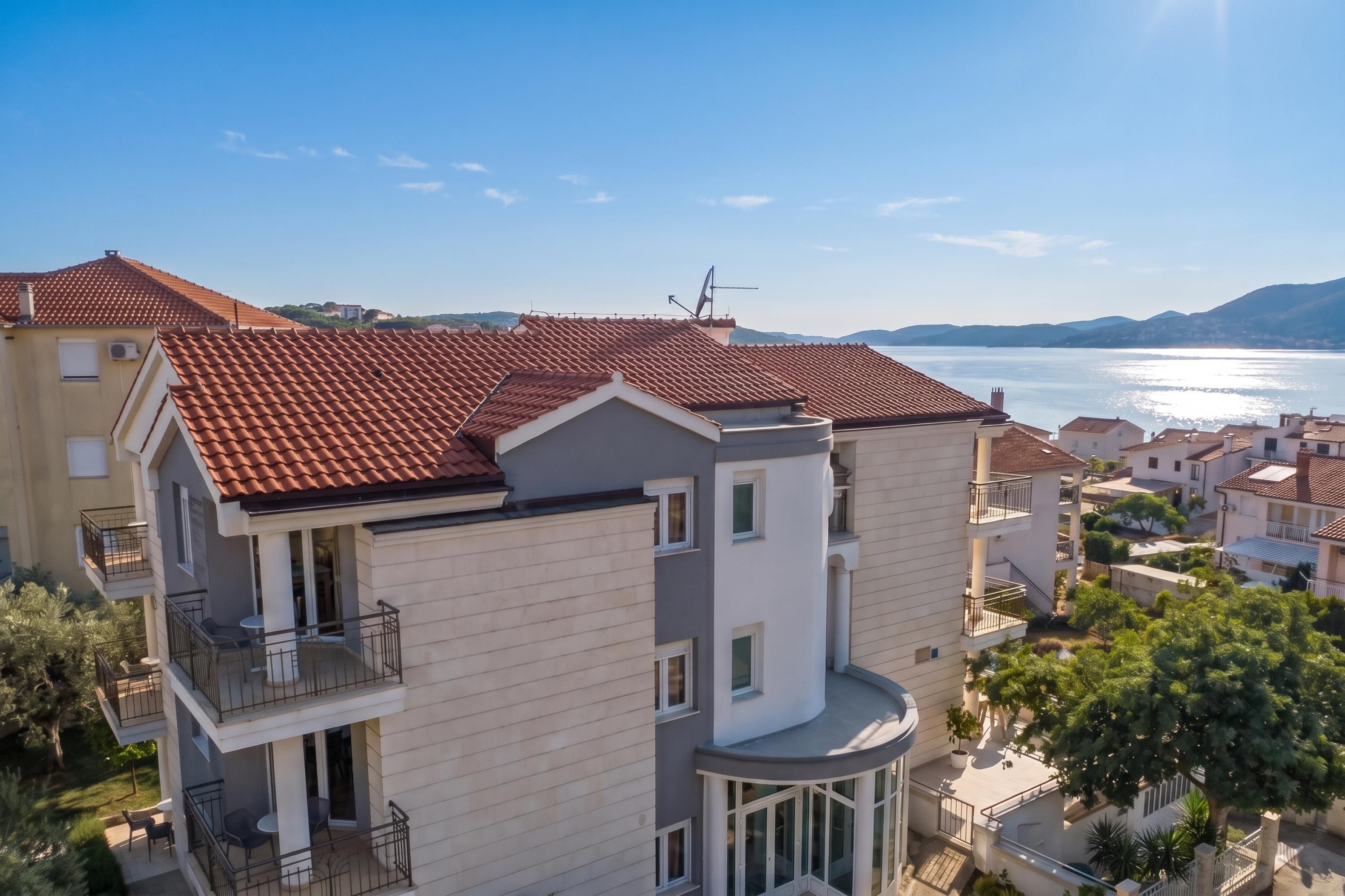 A multi-story residential building with a red-tiled roof, balconies, and a view of a body of water.