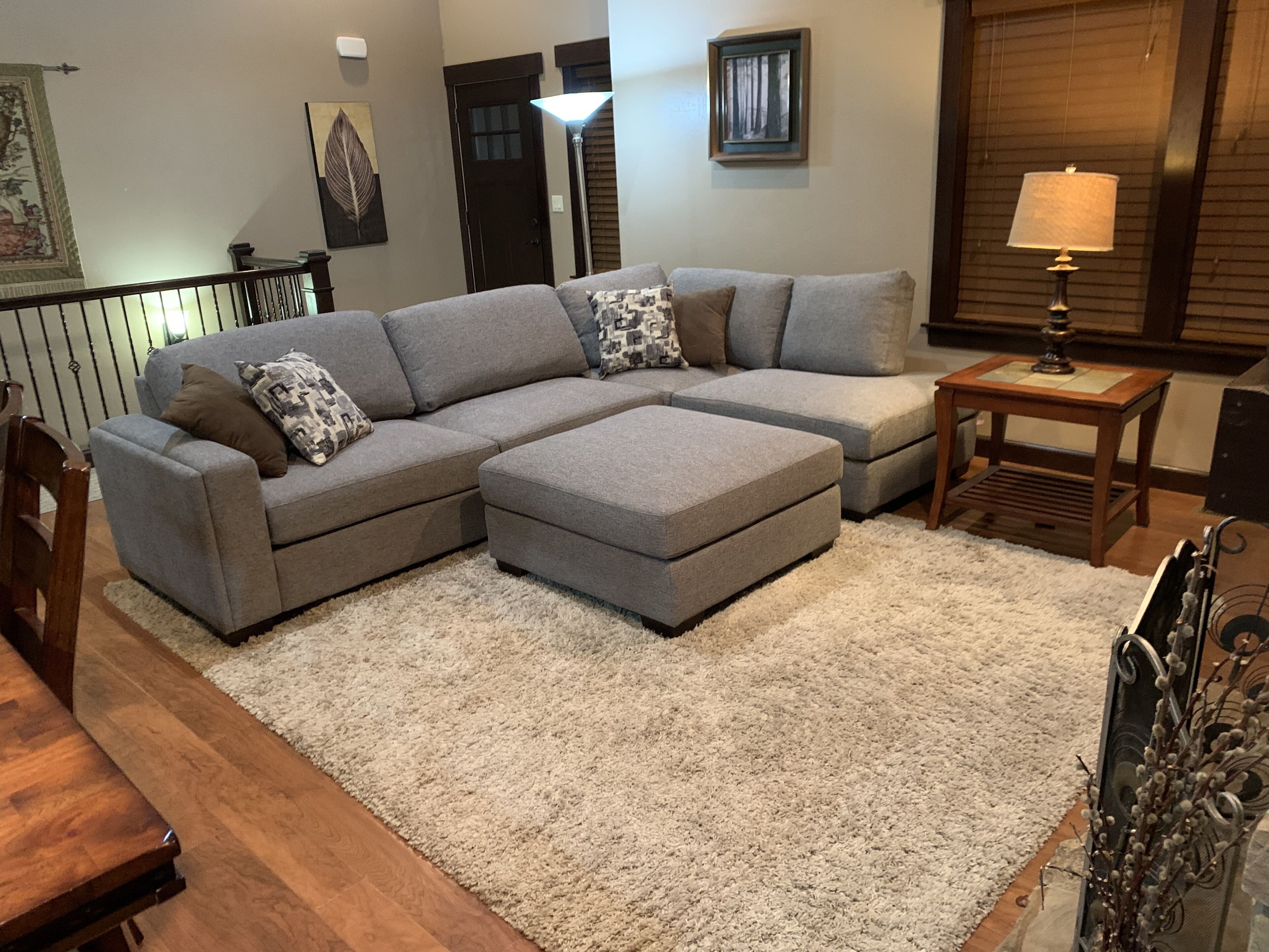 Ein Wohnzimmer mit einem grauen Ecksofa, einem kleinen Ottomanen, einem Holztisch und einem weißen Shaggy-Teppich.