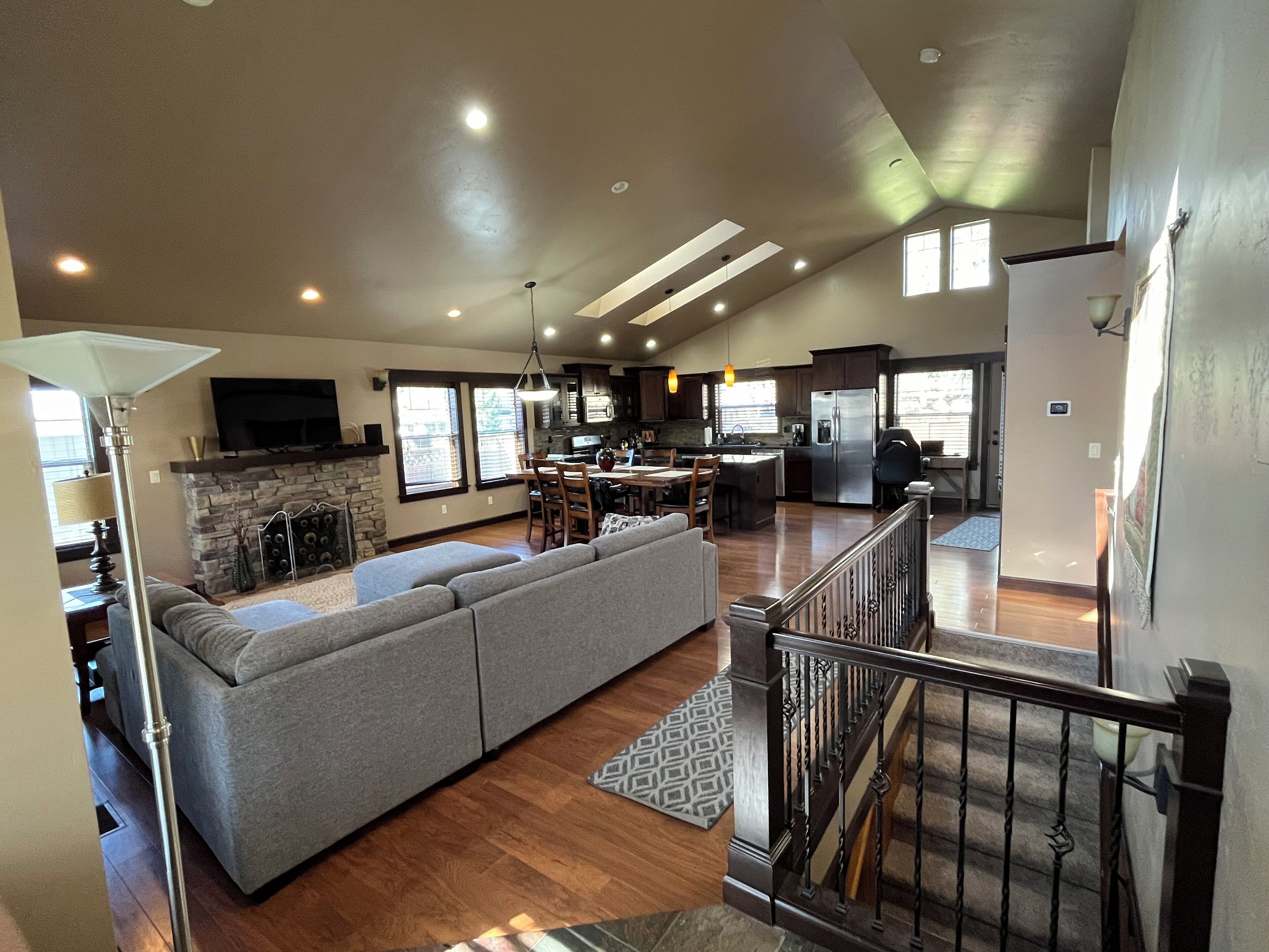 A spacious living area with a large gray sectional sofa, a fireplace, a dining table with chairs, and a kitchen in the background.