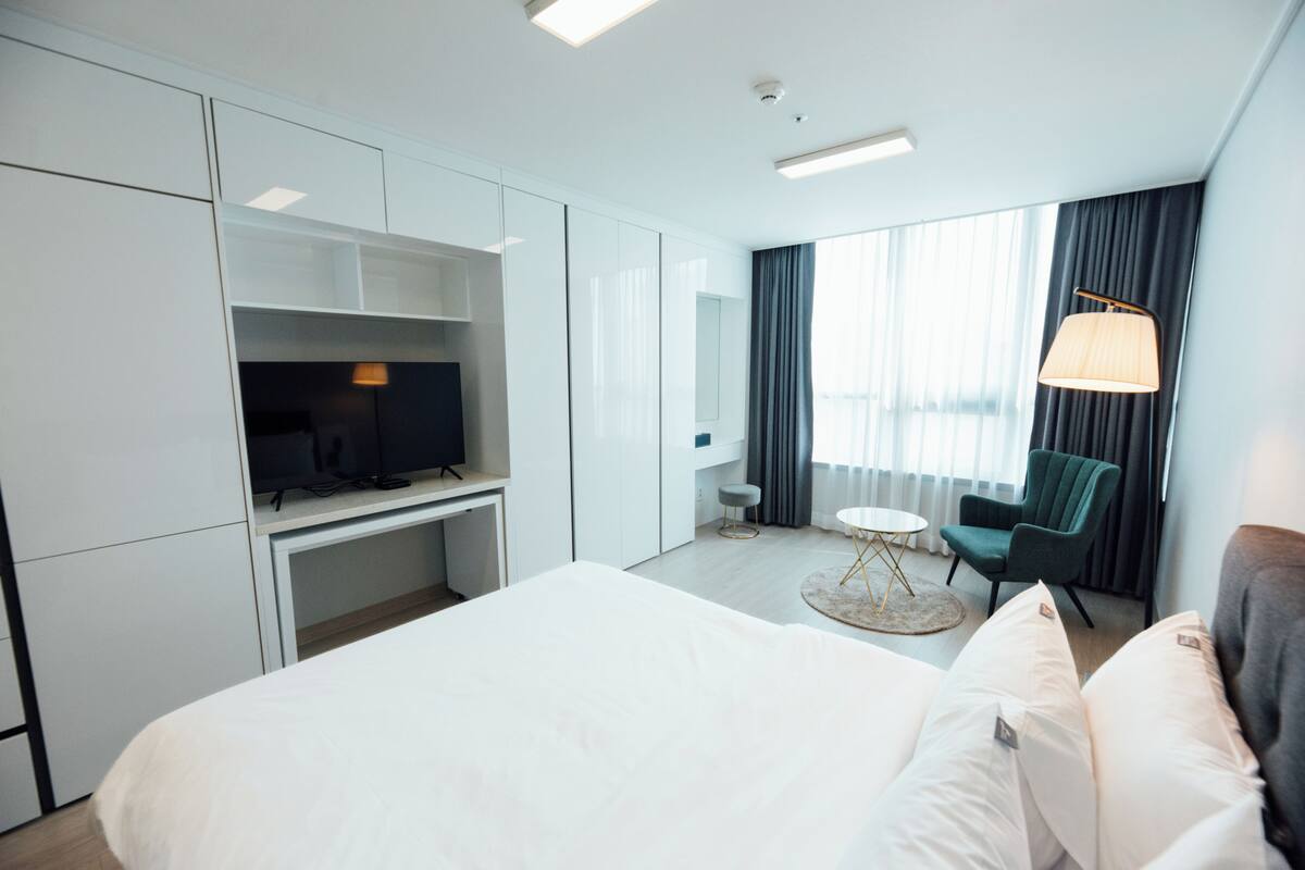 Habitación de hotel con cama, escritorio, televisor, silla, lámpara y ventana con cortinas.
