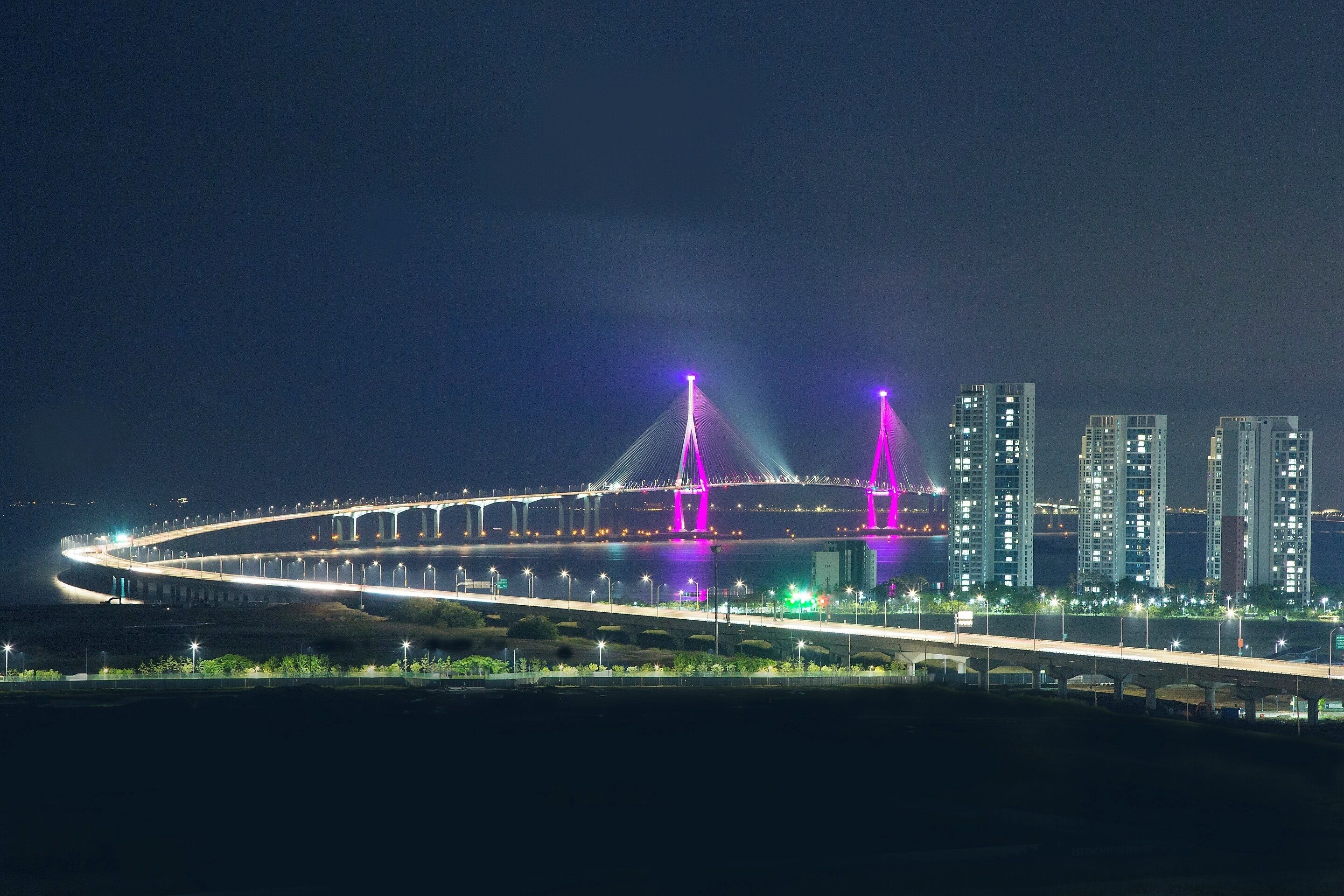 Vista nocturna de un puente moderno con estructuras iluminadas, silueta urbana de fondo y una carretera bien iluminada.