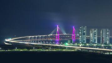 Vista nocturna de un puente moderno con estructuras iluminadas, silueta urbana de fondo y una carretera bien iluminada.