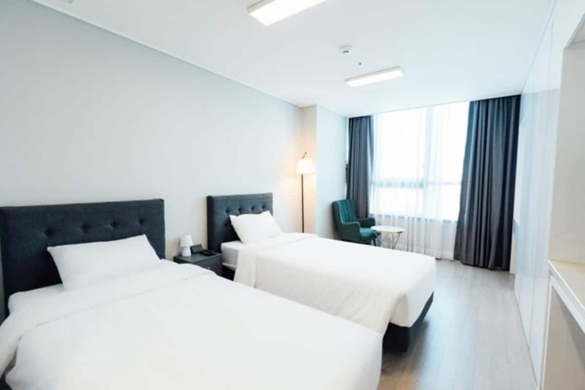 Habitación de hotel con dos camas, una mesita de noche, una lámpara, una silla y una ventana con cortinas.