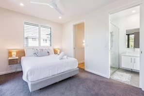 Quarto moderno com cama, luminárias de cabeceira, ventilador de teto, closet espaçoso e banheiro com box de vidro.