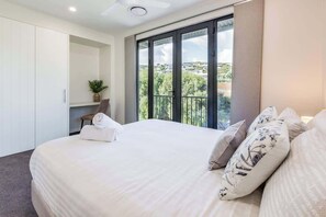 Een slaapkamer met een groot bed, een kledingkast, een zithoek met een plant en een balkon met uitzicht op de stad.