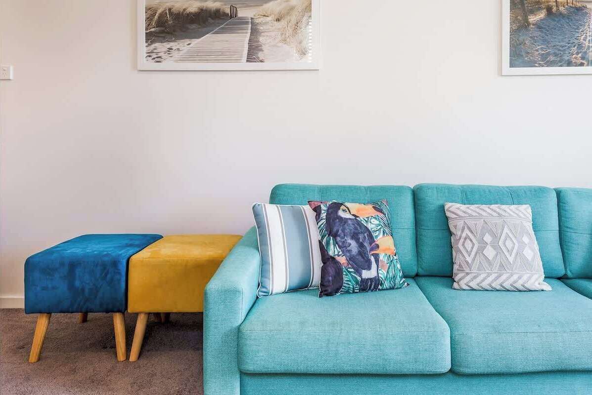A teal sofa with patterned cushions, a small yellow ottoman, and a framed picture on the wall.