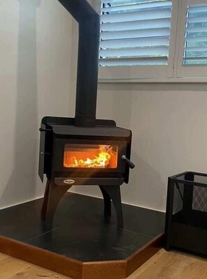 A wood-burning stove with a glass front, a black chimney, and a mesh basket to the right. - Yallingup Lagoon Beach House—A Spectacular Getaway (Yallingup)