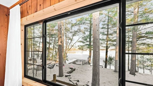 Une cabane dotée d’une grande fenêtre donnant sur une plage enneigée, bordée d’arbres, et possédant une terrasse en bois.