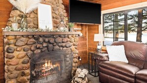 Une pièce de style chalet avec une cheminée en pierre où brûle un feu, un téléviseur fixé au mur et un canapé en cuir, le tout agrémenté de murs en bois et d’une fenêtre.