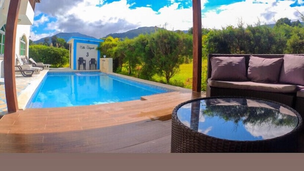 Una piscina con vistas a montañas y vegetación, un área de descanso con un sofá, y un edificio pequeño identificado como "Villa El Pescador 3".