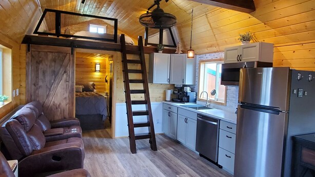 Ein gemütliches Blockhaus-Interieur mit Holzbalkendecke, einem Schlafzimmer auf der Empore, einer Küche mit Edelstahlgeräten und einem Ledersofa.