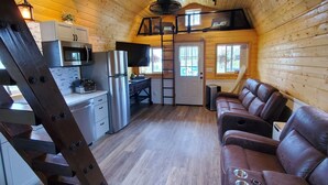 Ein gemütliches Blockhauskamin-Interieur mit Küche, Essbereich mit brauner Ledersofa und Schlafzimmer in der Mansarde, erreichbar über eine Leiter.