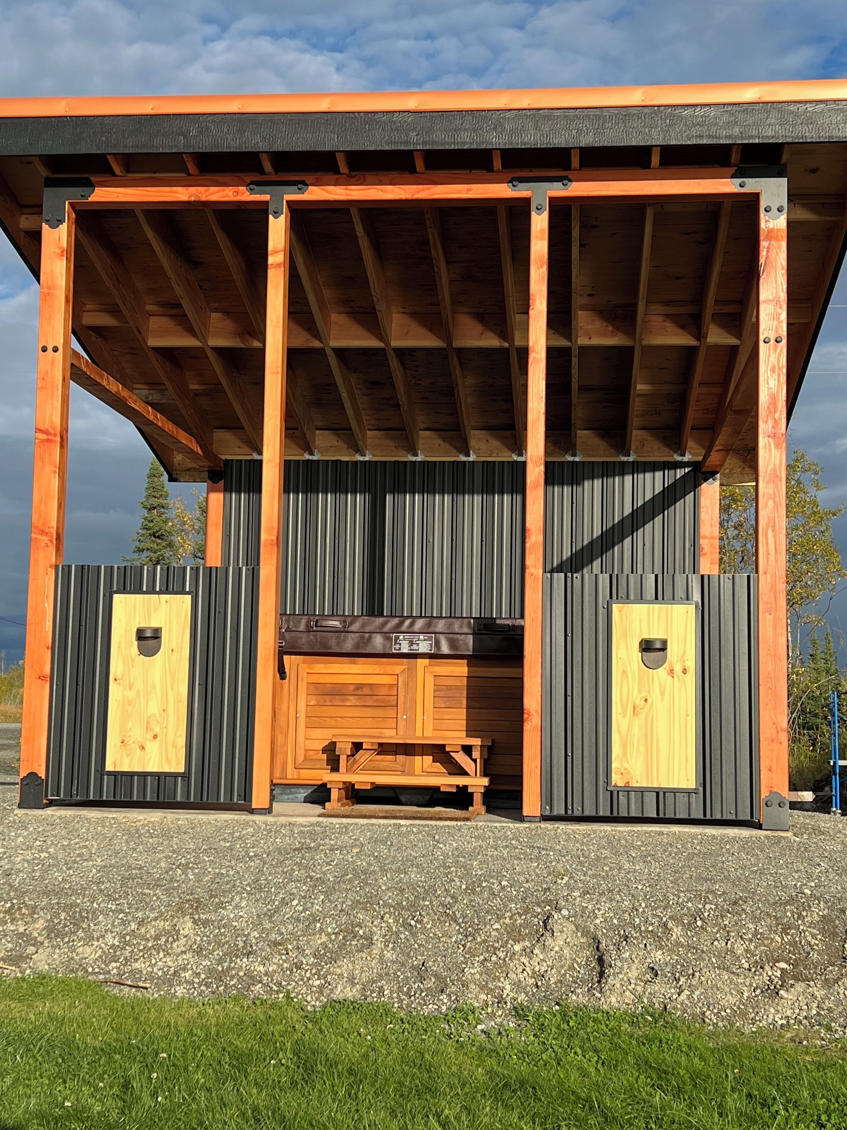 A covered outdoor area with a wooden bench and two doors, surrounded by metal siding and a gravel ground.