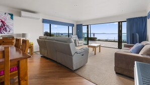 A modern living room with a grey sofa, a wooden dining table, and a view of the sea through large windows. - Waitetoko Waterfront - Waitetoko Holiday Home (Waitetoko)