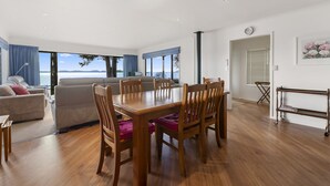 A modern living room with a wooden dining table, comfortable seating, and large windows offering a view of the sea. - Waitetoko Waterfront - Waitetoko Holiday Home (Waitetoko)
