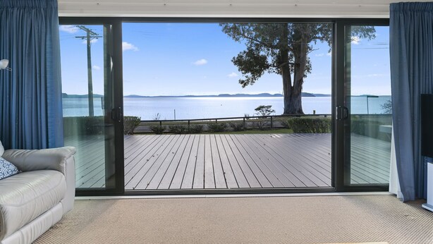 A room with a large sliding glass door leading to a wooden deck overlooking a body of water and distant land. - Waitetoko Waterfront - Waitetoko Holiday Home (Waitetoko)