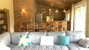 A cozy living room with a gray sectional sofa, wooden dining table, and chairs. A kitchen area is visible in the background with modern appliances and pendant lights.