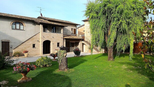 Una casa de dos plantas con fachada de piedra, tejado de tejas y un jardín cuidado con césped, un árbol y macetas.