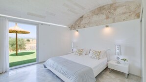 A bedroom with a large bed, a nightstand, a lamp, and a view of a grassy area through a sliding door. - Holiday Home "Es Gassons" with Pool, Wi-Fi, AC, Terrace & Garden (María de la Salut)