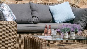 A wicker sofa with cushions, a glass coffee table with bottles and plants, and a stone wall in the background. - Holiday Home "Es Gassons" with Pool, Wi-Fi, AC, Terrace & Garden (María de la Salut)