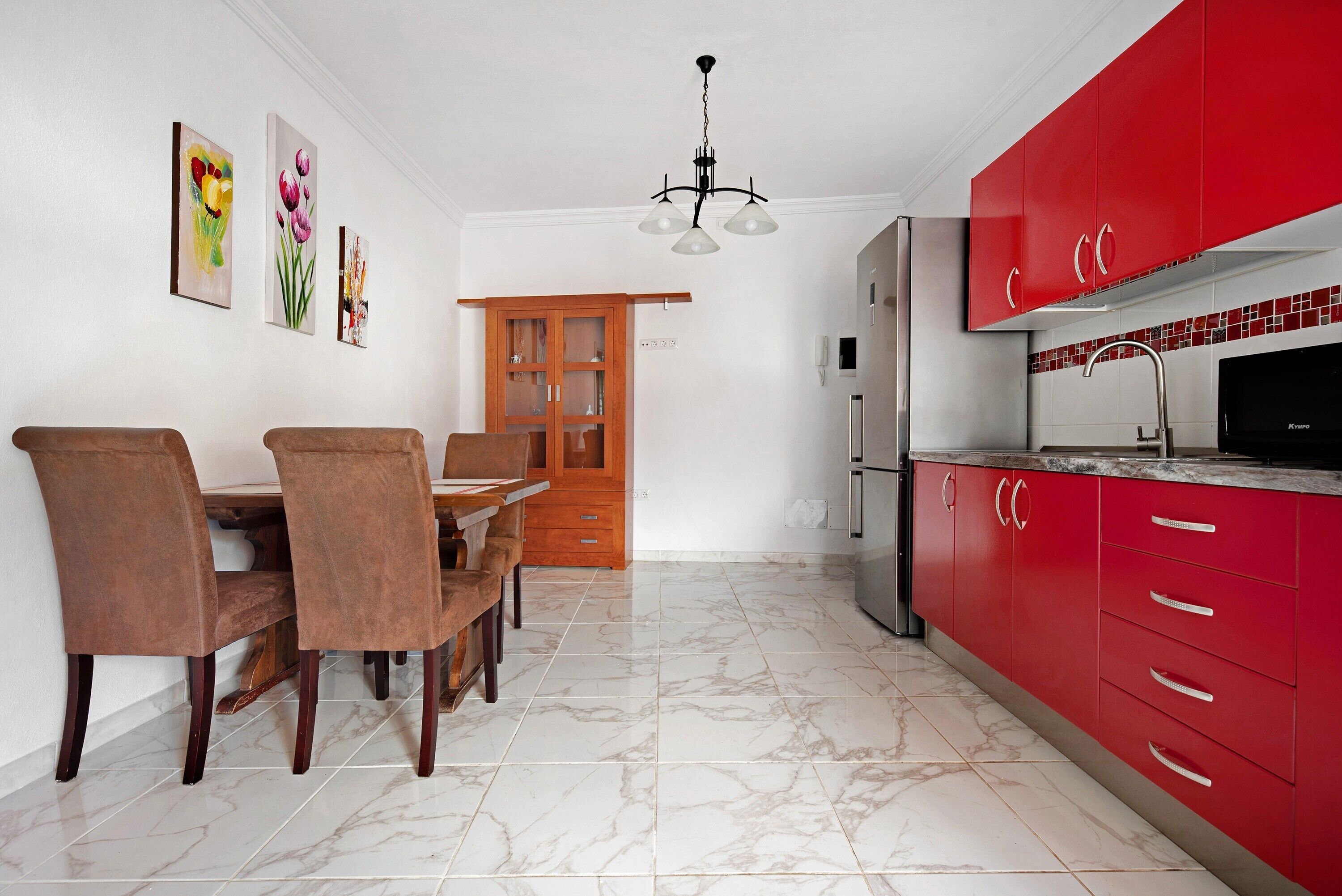 Una cocina con armarios rojos, suelo de mármol y zona de comedor con sillas y una mesa.