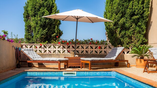 A pool area with lounge chairs and an umbrella, surrounded by a decorative fence and greenery. - Holiday Home "Villa Sion" with Mountain Views, Pool, Wi-Fi & Terrace, Near Beaches (Cala Mendia)