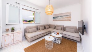 A modern living room with a sectional sofa, a white cabinet, a large window, a hanging light fixture, and a mounted TV. - Holiday Home "Villa Sion" with Mountain Views, Pool, Wi-Fi & Terrace, Near Beaches (Cala Mendia)