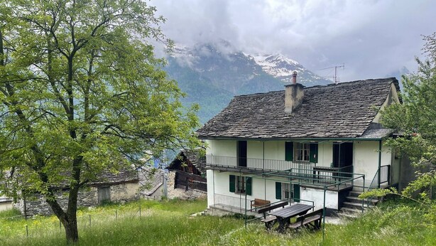 Une maison à deux étages dotée d’un balcon, entourée d’une végétation luxuriante et ayant une chaîne de montagnes en toile de fond.