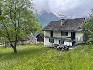 Ein zweistöckiges Haus mit Balkon, umgeben von üppiger Vegetation und einer Bergkulisse.