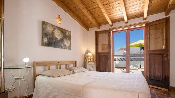 Une chambre avec un plafond en bois, un lit recouvert de draps blancs, une table de chevet avec une lampe et une grande fenêtre donnant sur l’extérieur.