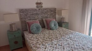 A bedroom with a stone-textured headboard, floral bedding, two nightstands with lamps, and a 'Welcome to the Beach' sign.