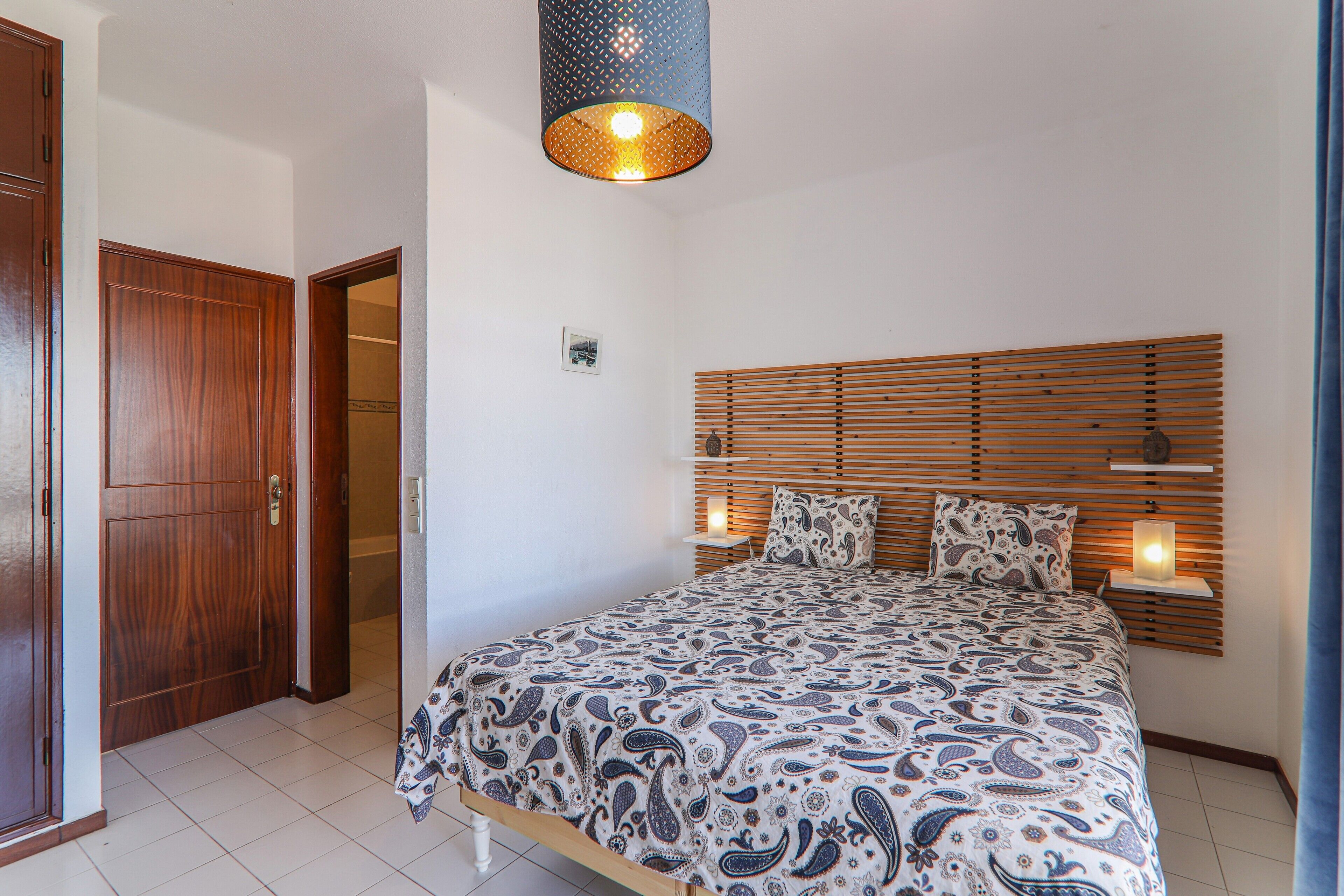 Een slaapkamer met een houten hoofdbord, een bed met een decoratieve spreidekker, een bedlampje en een deur.