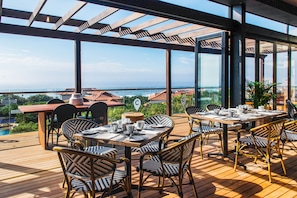 An outdoor dining area with tables and chairs, overlooking a scenic view of the ocean and buildings. - The Capital Zimbali (Ballito)