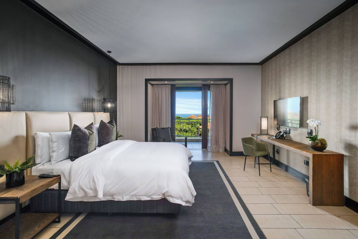 A modern hotel room with a large bed, a desk, a chair, a TV, and a balcony with a view.