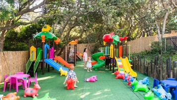 A playground with slides and climbing structures, surrounded by a wooden fence and trees.