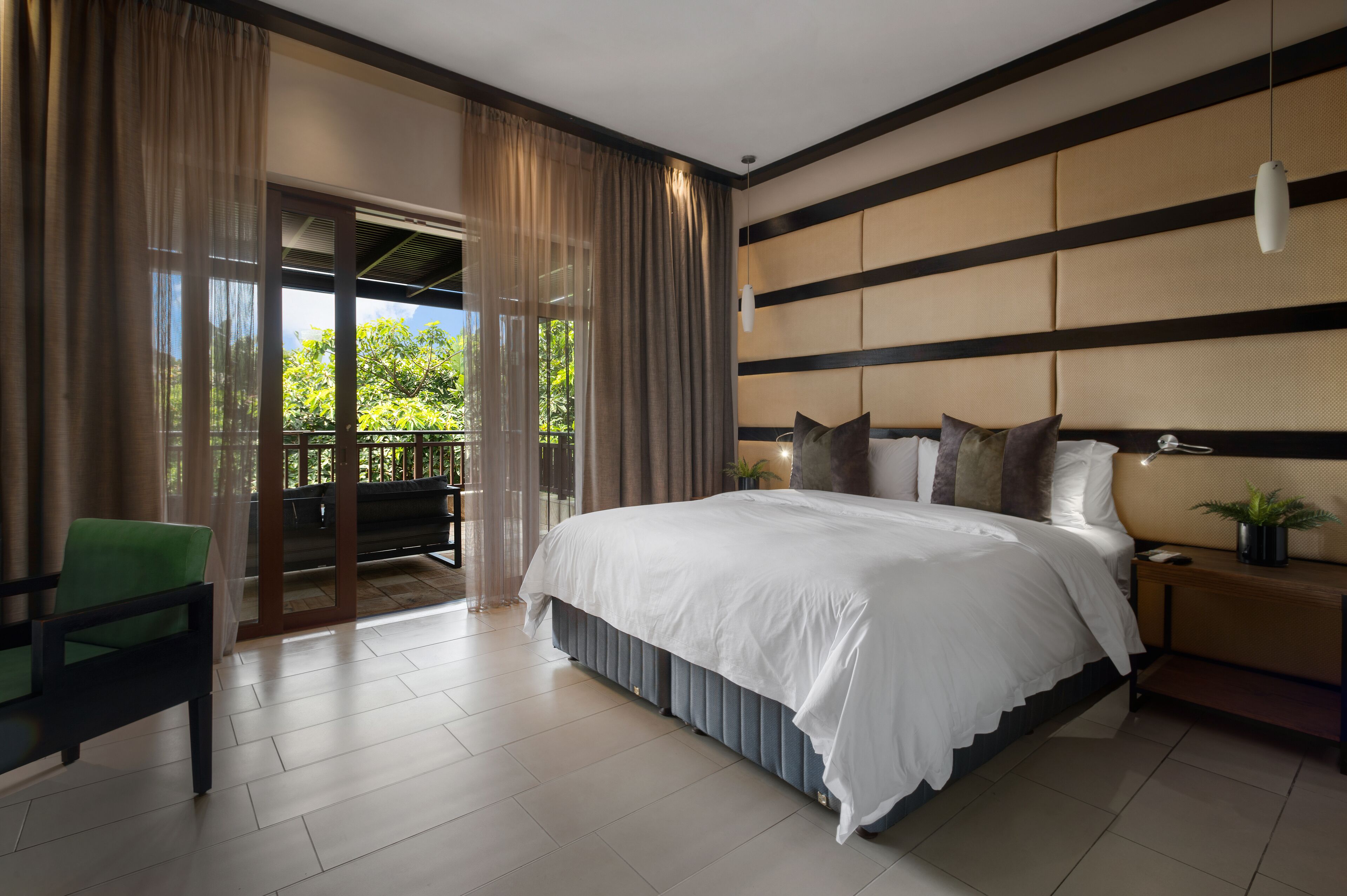 A spacious bedroom with a large bed, a nightstand, a green chair, and a view of greenery through the window.