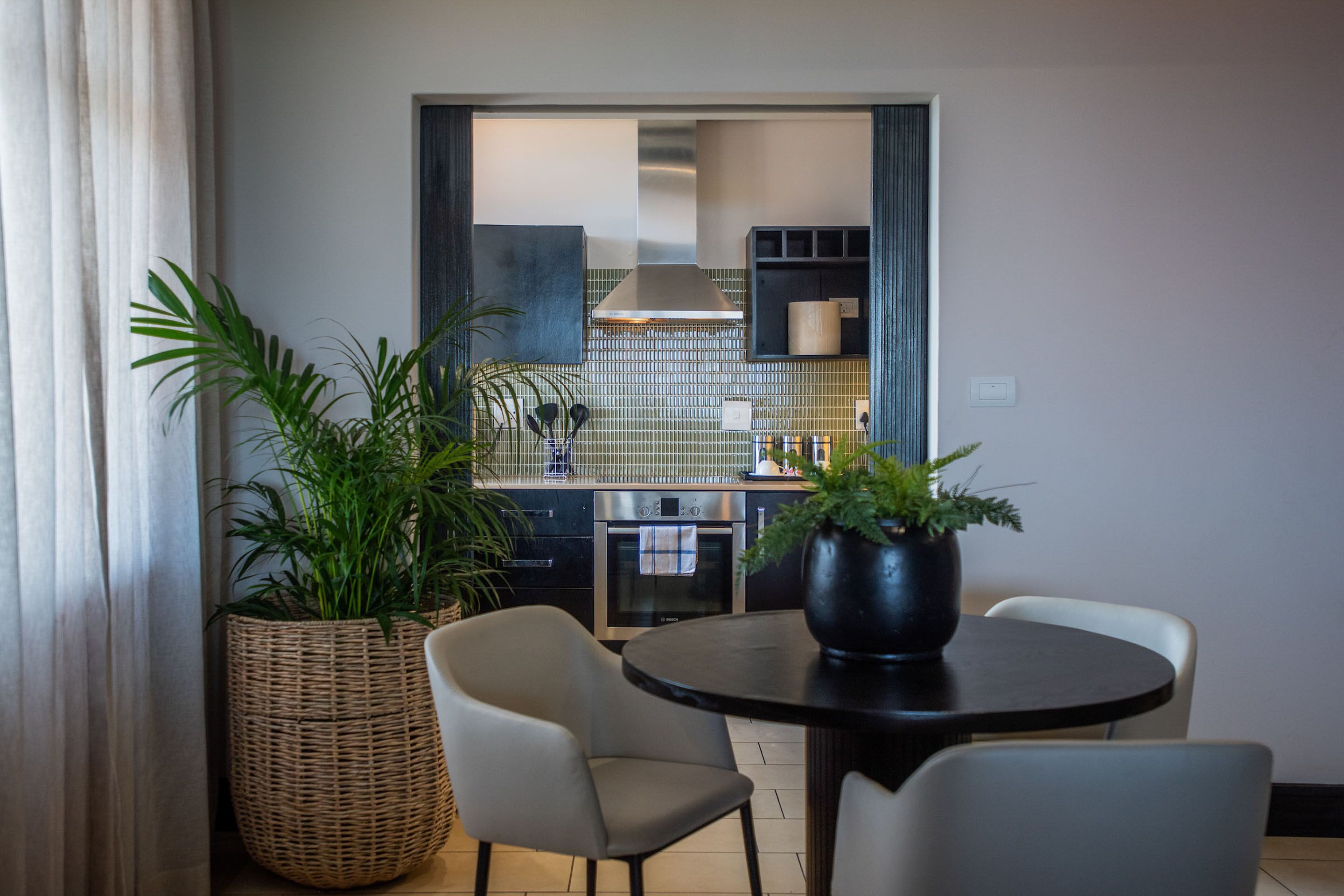 A dining area with a round table and two chairs, a large woven basket with a plant, and a view into the kitchen with a tiled backsplash and stainless steel appliances.