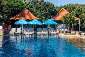 A swimming pool with lounge chairs and blue umbrellas, surrounded by trees and a red-roofed building. - The Capital Zimbali (Ballito)