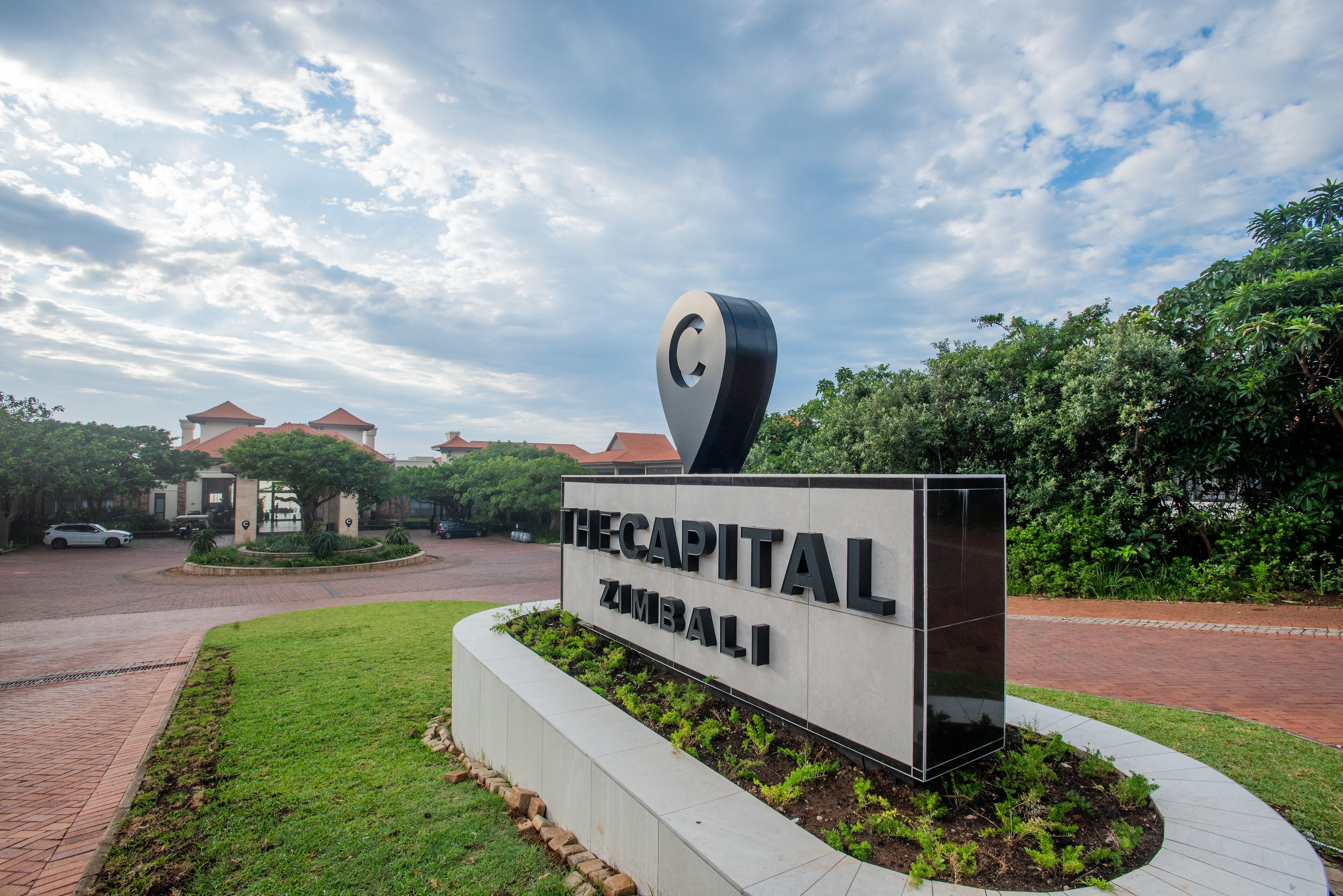 Un cartel para 'The Capital Zimbali' con un diseño moderno, rodeado de un área ajardinada con césped y arbustos.